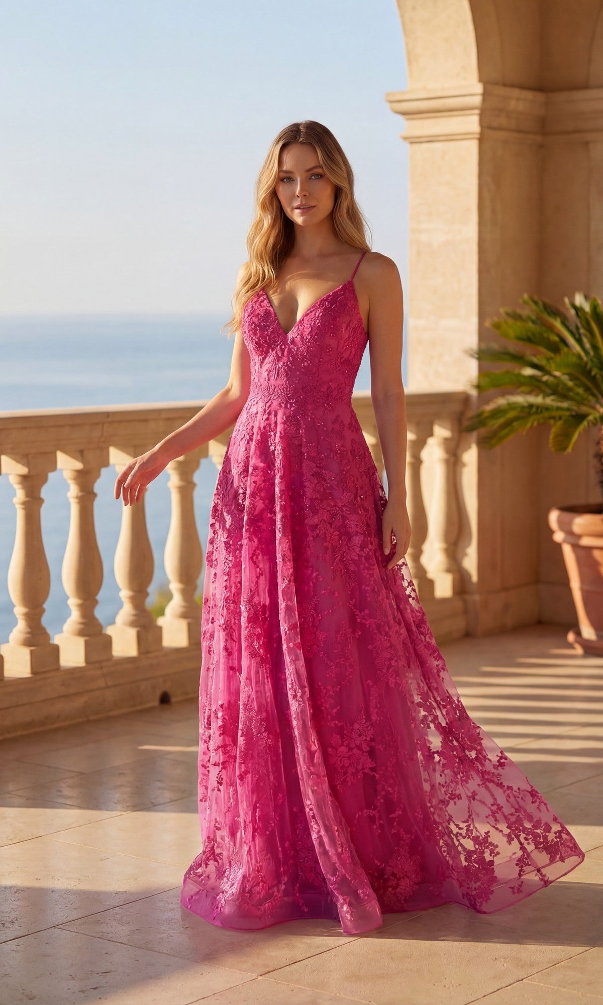 A woman in the Long Prom Dress: Chicas C804, a sleeveless floor-length gown in fuchsia, with floral lace embroidery, stands on a sunlit balcony overlooking the coast, framed by elegant stone columns.